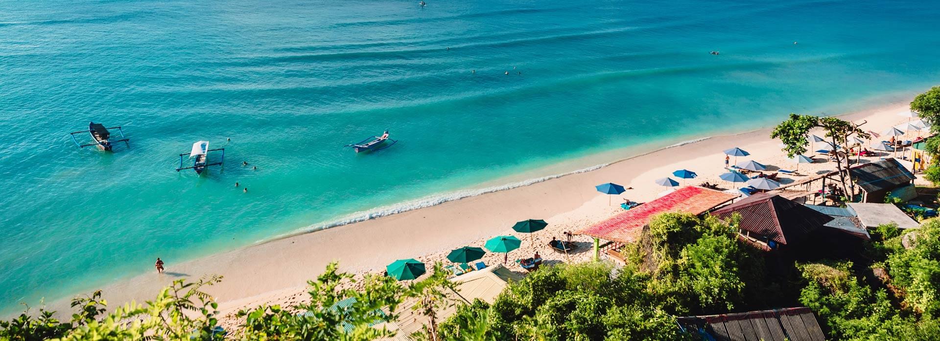 A beach with turquoise water, white sand, and green trees, with several boats in the water and umbrellas and lounge chairs on the sand.